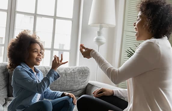 African disabled daughter and her mother sitting on sofa show symbols with hands using visual-manual finger gestures interacting at home. 
