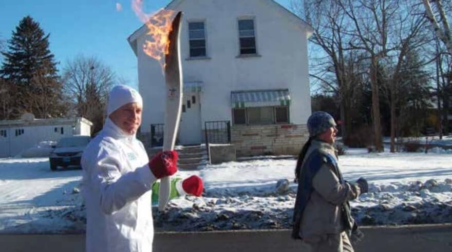 A man carrying the Olympic torch in front of a house with a snowy yard
